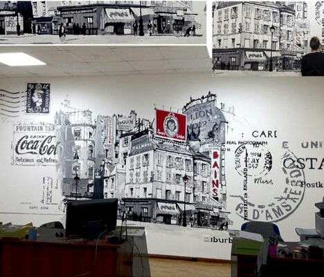 Ściana biura ozdobiona czarno-białym malowidłem przedstawiającym ulicę z budynkami w stylu retro, uzupełnionym o elementy graficzne przypominające stare pocztówki i reklamy, nad biurkiem z monitorem.