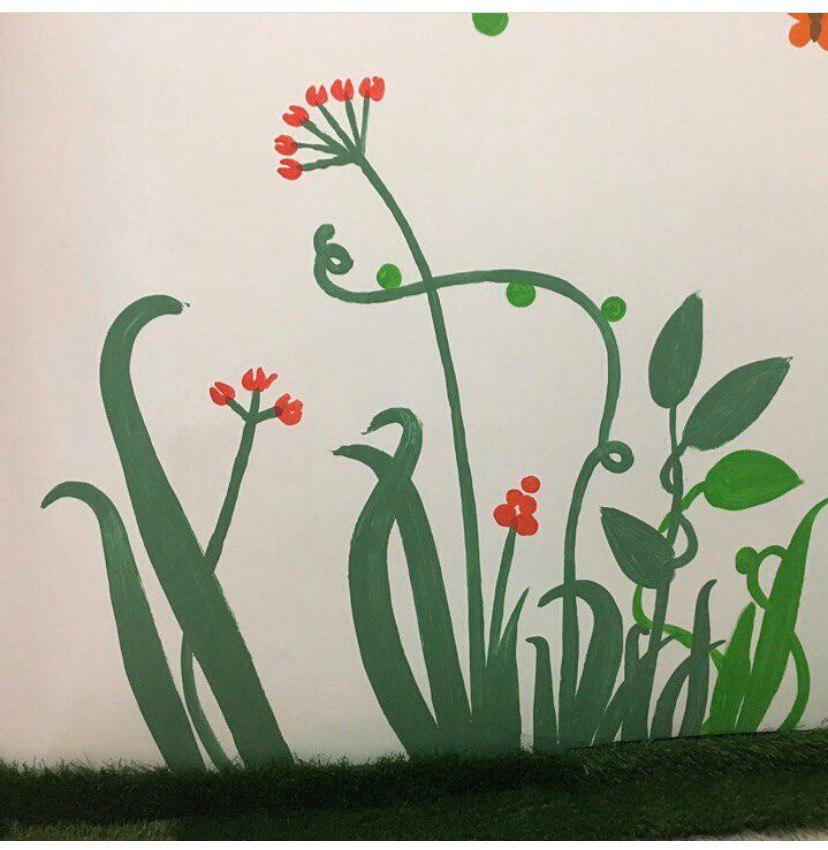 Ręcznie malowana, dziecięca dekoracja ścienna z motywem stylizowanych roślin i czerwonych kwiatów na białym tle, z imitacją trawy u dołu.