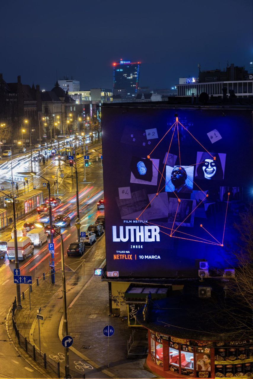 Wielkoformatowy billboard reklamowy filmu Netflix 'Luther: Zmrok' nocą, widoczny z perspektywy ulicy z ruchem samochodowym i światłami miasta w tle.