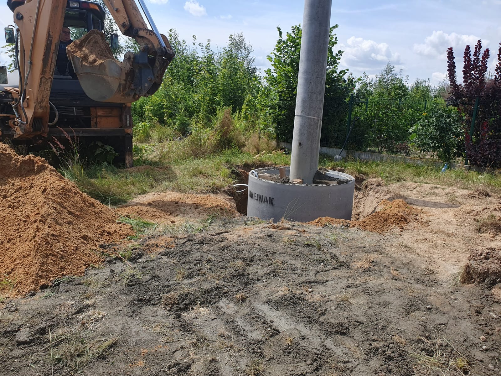 Żółta koparka podczas prac ziemnych przy betonowym kręgu z zamontowanym słupem, widoczne ślady opon na ziemi i usypana pryzma piasku.
