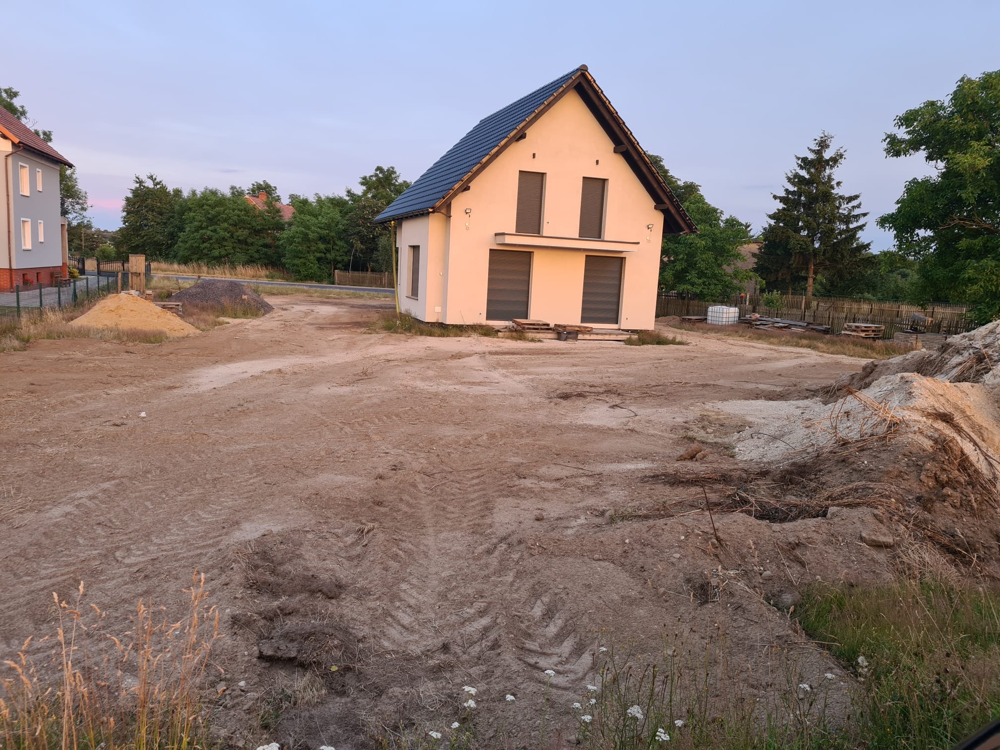 Nowo wybudowany, parterowy dom z ciemną dachówką, stojący na wyrównanym, piaszczystym terenie z widocznymi śladami opon, w tle drzewa i zabudowania.