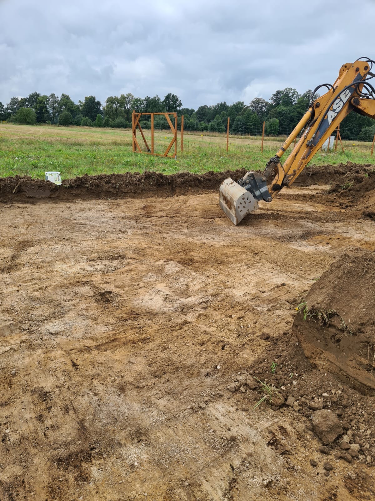 Żółta koparka CASE w trakcie prac ziemnych, widoczny wykop z równą krawędzią, w tle drewniana konstrukcja i pole pod pochmurnym niebem.