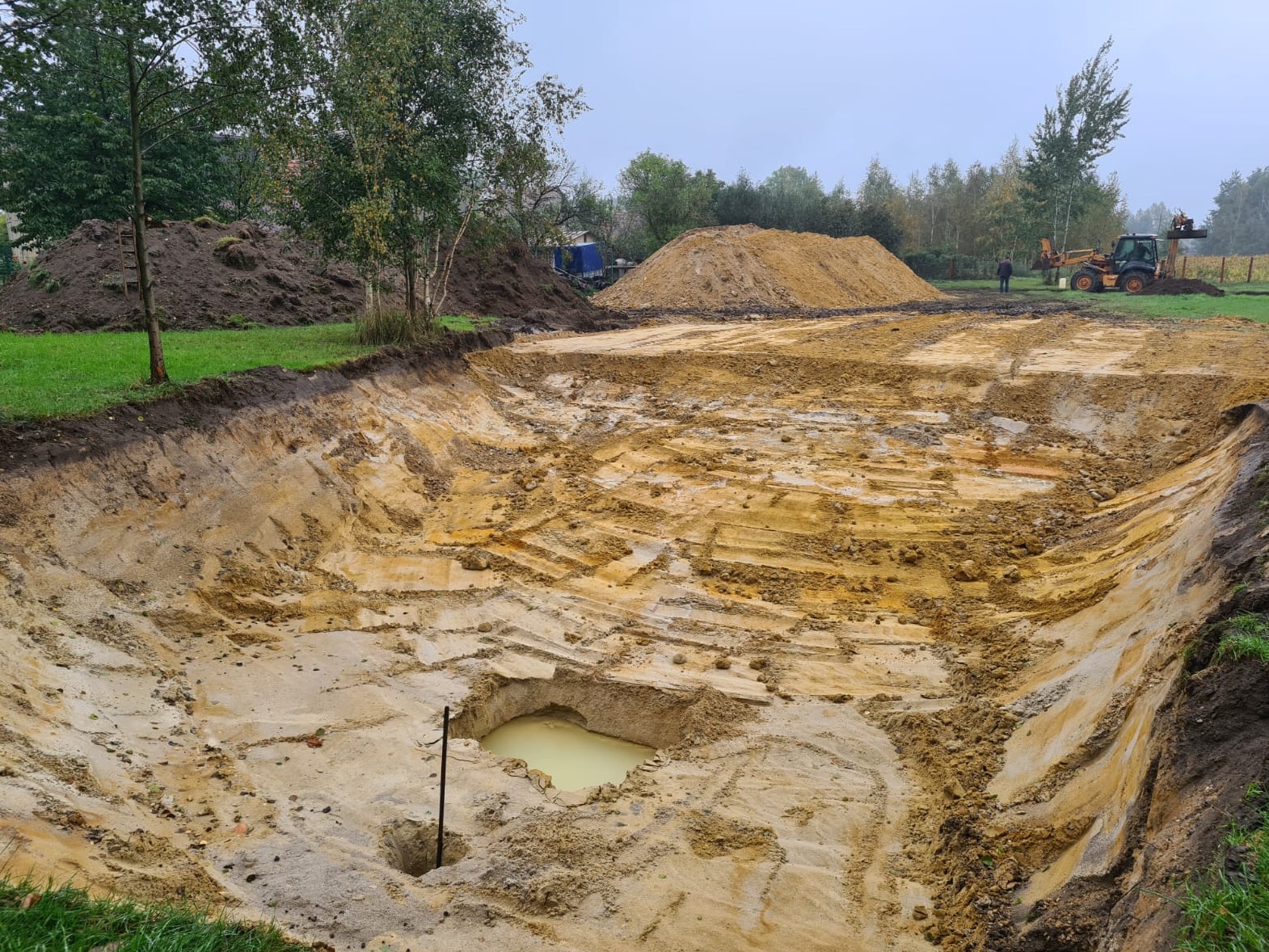 Szeroki wykop w ziemi z widocznymi śladami opon, mała sadzawka z wodą w dole wykopu, w tle hałdy ziemi i maszyna budowlana w oddali.