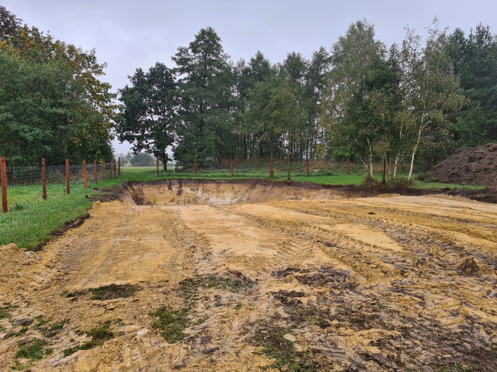 Wykop pod staw w trakcie realizacji, widoczne ślady opon na piaszczystym dnie, ogrodzenie z siatki leśnej i drzewa w tle, pochmurny dzień.
