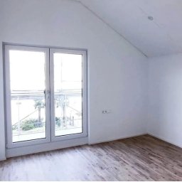 RemontNaJuż - Jasne wnętrze poddasza z drewnopodobną podłogą, białymi ścianami i oknem balkonowym z widokiem na zieleń.