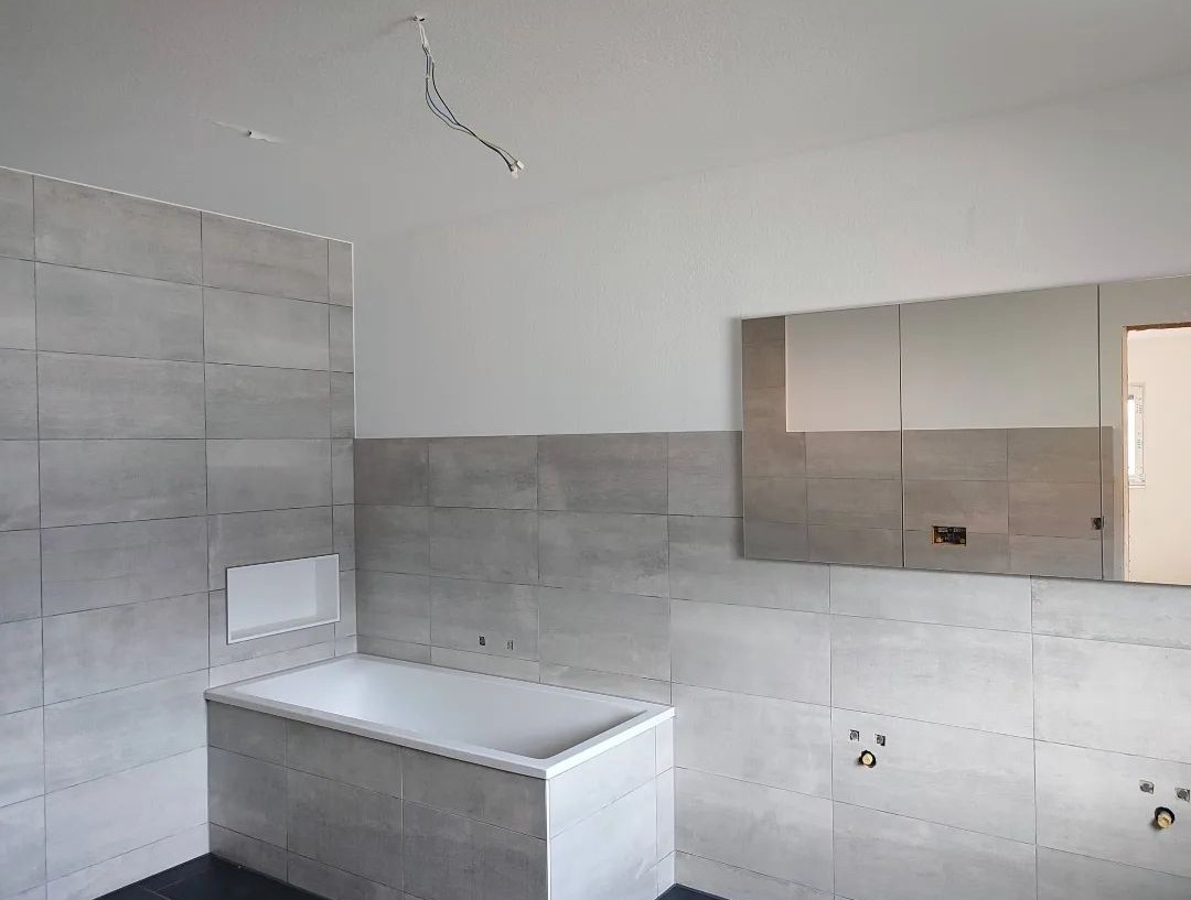 Łazienka w trakcie wykończenia z szarymi płytkami, białą wanną z wnęką, lustrem i wystającymi przewodami elektrycznymi z sufitu.