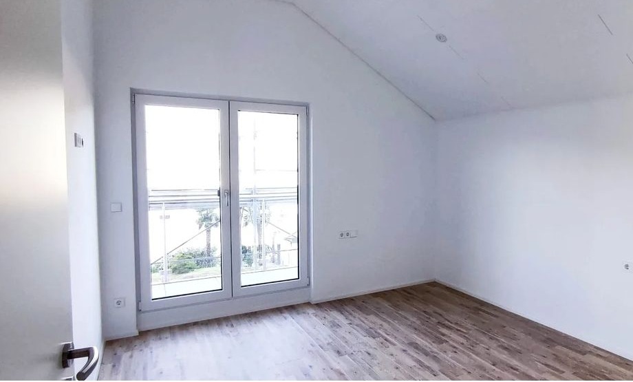 Jasne wnętrze poddasza z drewnopodobną podłogą, białymi ścianami i oknem balkonowym z widokiem na zieleń.