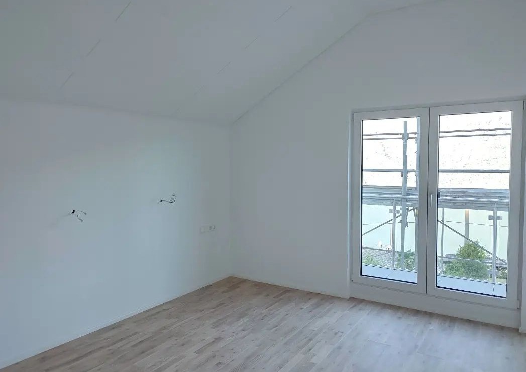 Wnętrze pokoju z jasnymi panelami podłogowymi, białe ściany z wystającymi kablami, dwuskrzydłowe okno z widokiem na rusztowanie i zieleń na zewnątrz.