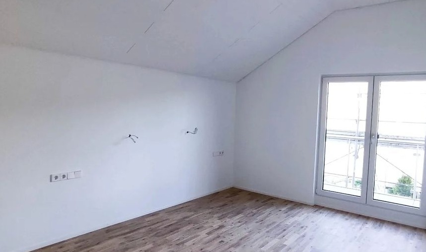 Pokój po remoncie z jasnymi ścianami, panele podłogowe w odcieniach szarości i biale okno z widokiem na zewnątrz; widoczne wystające kable i gniazdka elektryczne.