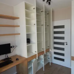 Biała szafa na wymiar z otwartymi drzwiami, drewniane półki i biurko z komputerem, minimalistyczny design w jasnym wnętrzu.