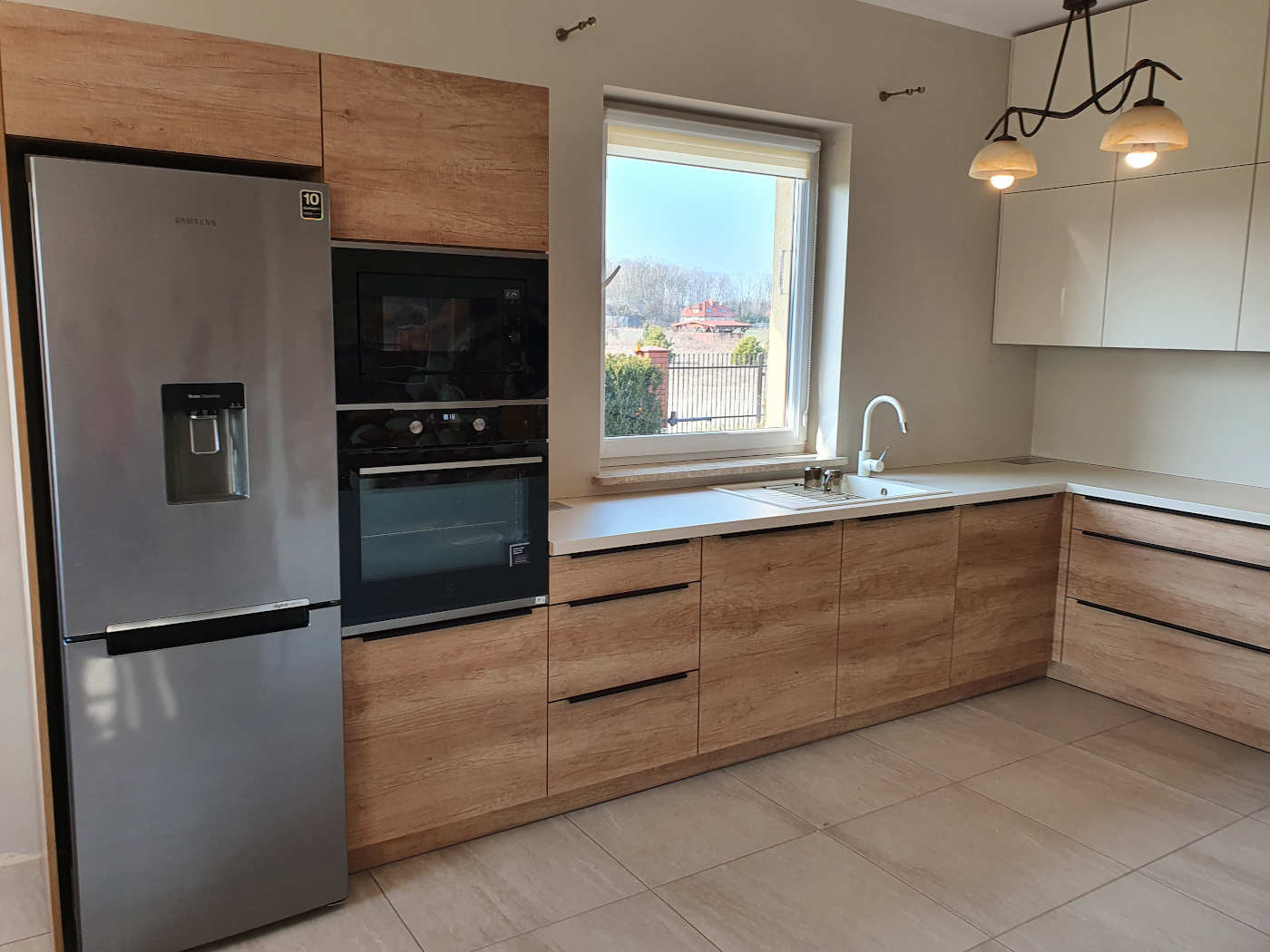 Jasna kuchnia z drewnopodobnymi frontami szafek, srebrną lodówką z dystrybutorem wody, białym zlewem pod oknem i trzema lampami w stylu retro.