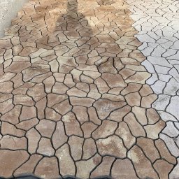 Maksym Kolisnichenko - Fragment nawierzchni z nieregularnych, beżowo-brązowych elementów, częściowo ułożonych, z widocznym cieniem osoby i przygotowanym podłożem z boku.