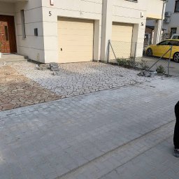 Maksym Kolisnichenko - Układanie kostki brukowej przed domem z dwoma garażami, widoczne dwa rodzaje kostki: szara ułożona i brązowa w trakcie układania, obok sterta kostki do ułożenia.