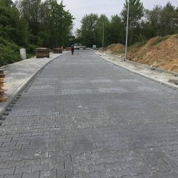 Maksym Kolisnichenko - Nowo ułożona, szara kostka brukowa w kształcie litery 'H' na długiej drodze; w tle widać pracownika, ciężarówkę i stosy palet z kostką.