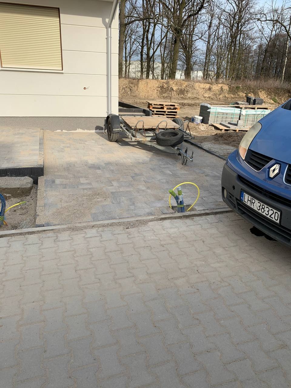 Fragment nowej nawierzchni z szarej kostki brukowej przed budynkiem z przyczepą i niebieskim samochodem, widoczne przygotowane materiały budowlane i piasek.
