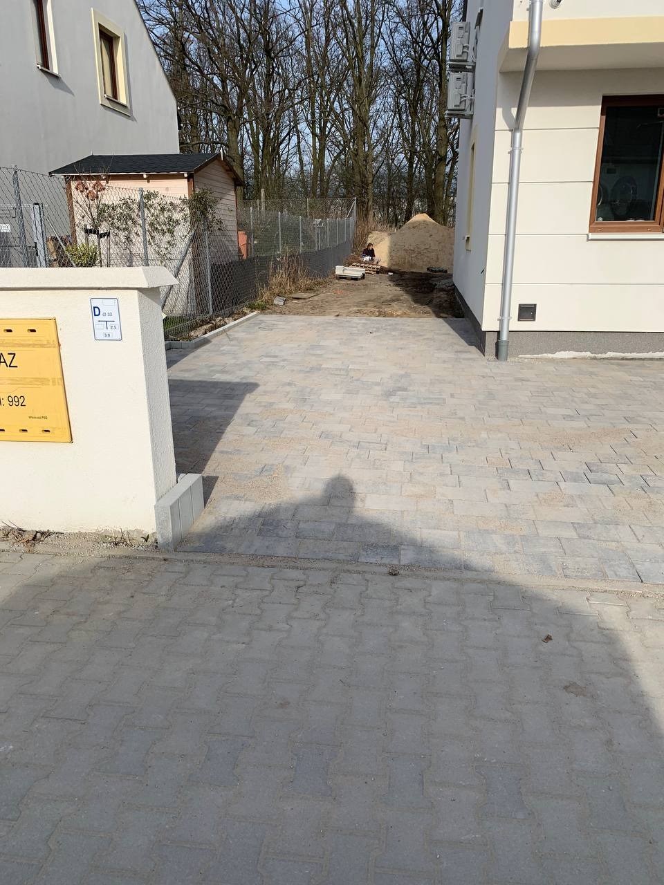 Nowo ułożona szara kostka brukowa na podjeździe, widoczny cień osoby robiącej zdjęcie, w tle budynek i sterta piasku.