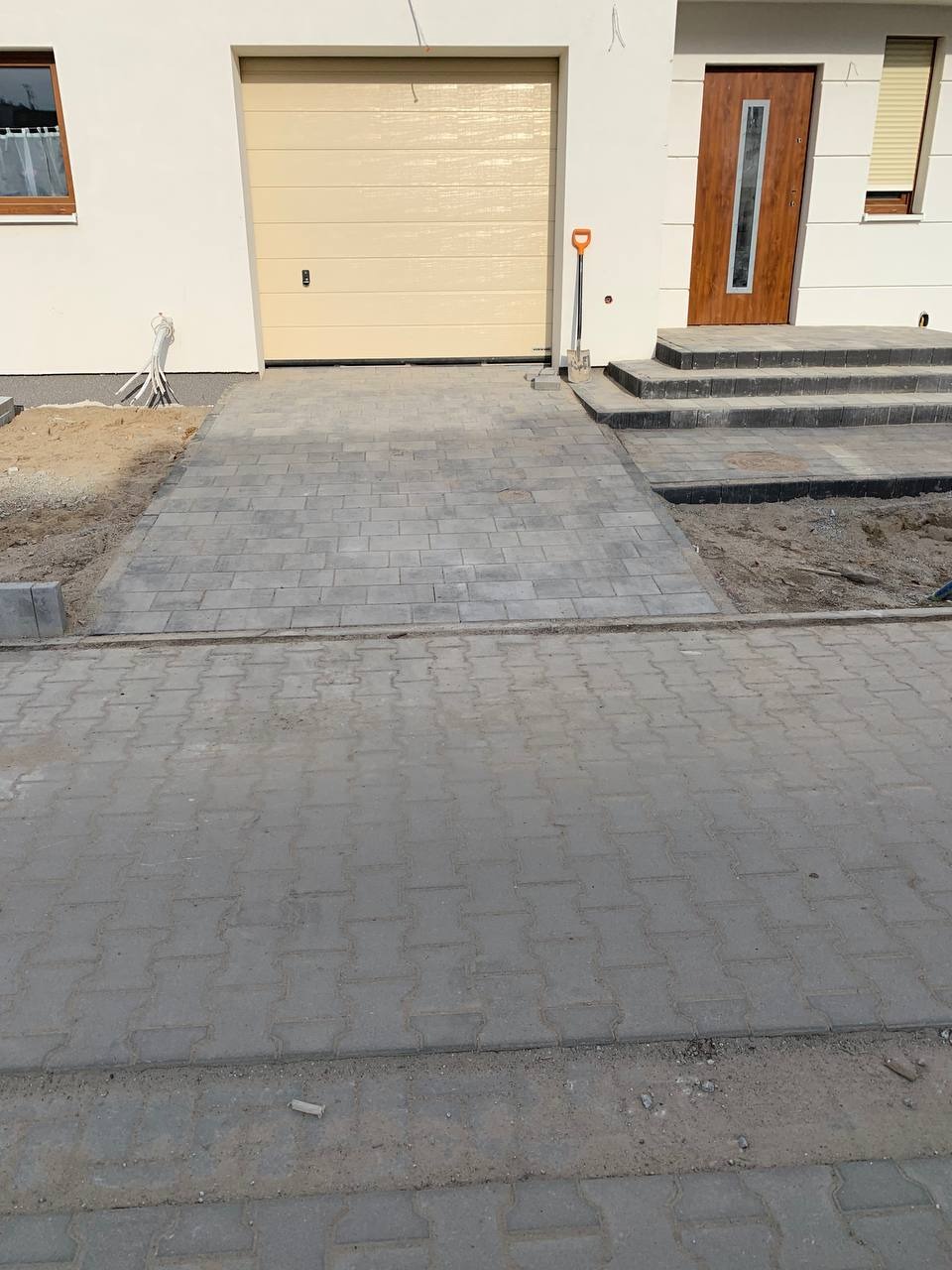 Szara kostka brukowa ułożona przed domem z beżową bramą garażową i brązowymi drzwiami wejściowymi, widoczna łopata przy schodach, fragmenty piasku i ziemi wskazują na trwające prace.