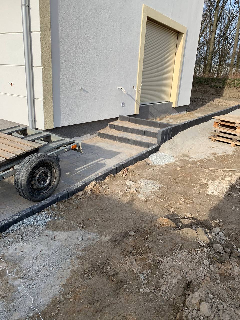 Układanie kostki brukowej wzdłuż ściany budynku z trzema schodami, widoczny fragment przyczepki i palety w tle, teren przygotowany pod dalsze prace.