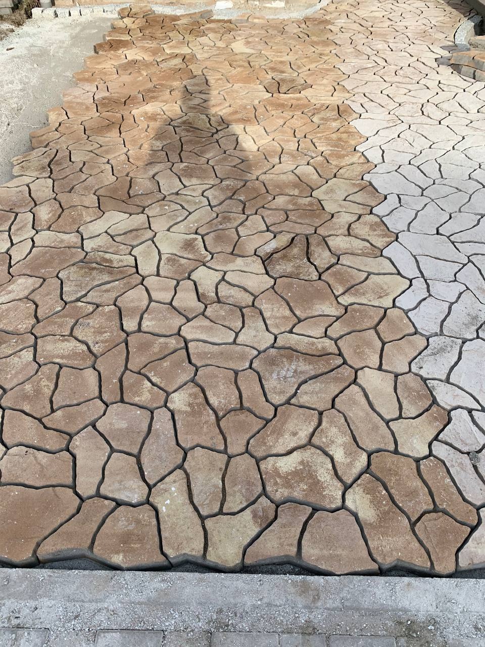 Fragment nawierzchni z nieregularnych, beżowo-brązowych elementów, częściowo ułożonych, z widocznym cieniem osoby i przygotowanym podłożem z boku.