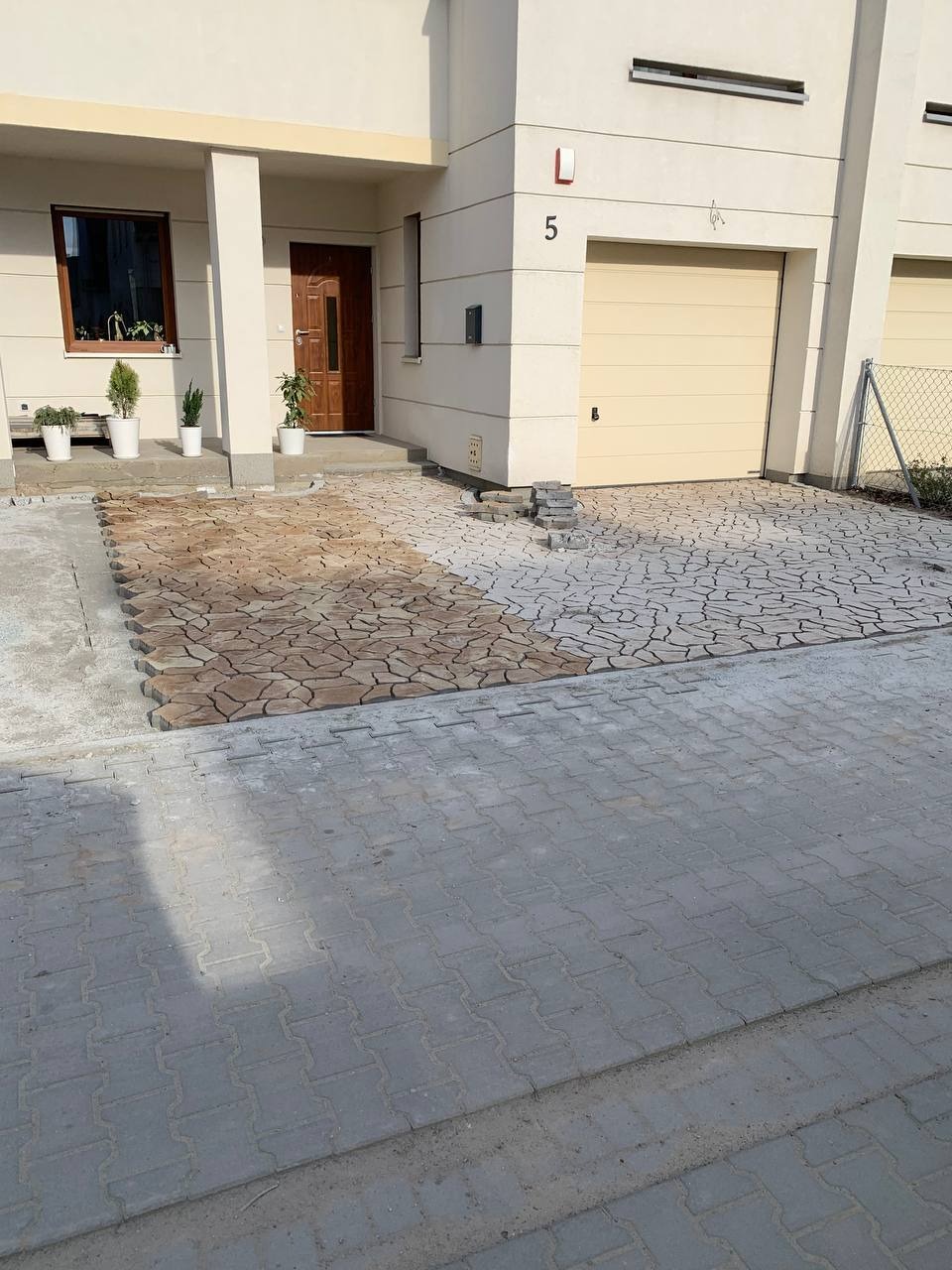 Fragment podjazdu przed domem jednorodzinnym, częściowo wyłożony szarą kostką brukową typu 'I', z obszarem przygotowanym pod ułożenie beżowej kostki o nieregularnym kształcie, widoczne palety...