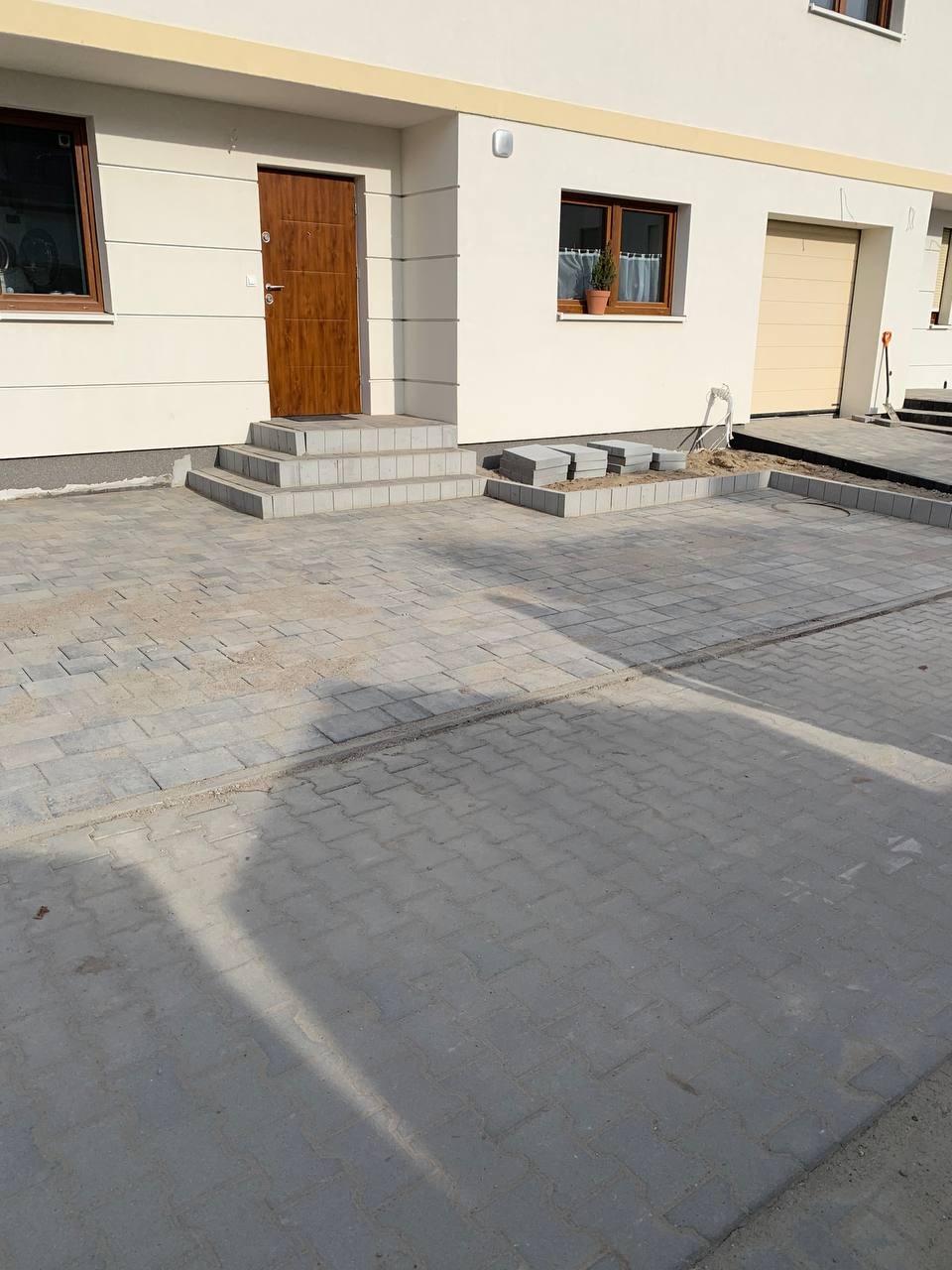 Ułożona szara kostka brukowa przed domem z brązowymi drzwiami i oknami, widoczne schody wejściowe i częściowo ułożona obwódka z kostki.