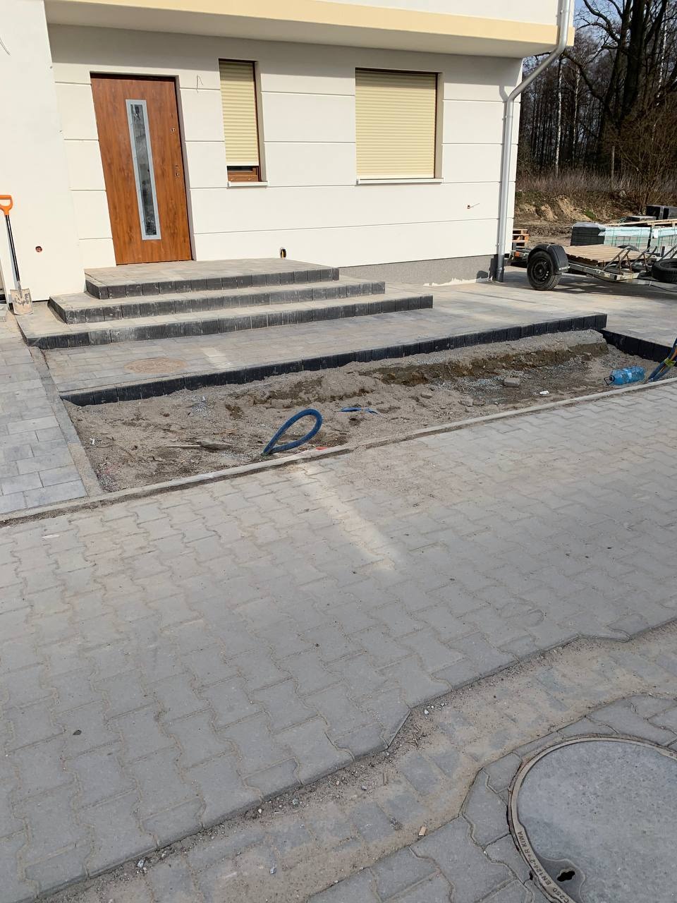 Przygotowanie terenu pod ułożenie nawierzchni z kostki, widoczny wykop obramowany kostką, częściowo ułożone schody wejściowe do budynku, niebieski wąż na piasku.