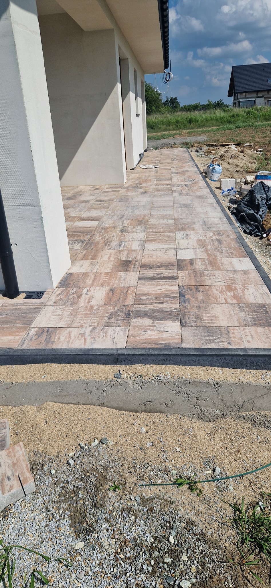 Ułożona kostka brukowa imitująca drewno, obrzeża z ciemnego kamienia, widoczna podbudowa z piasku i betonu, fragment elewacji budynku w tle.