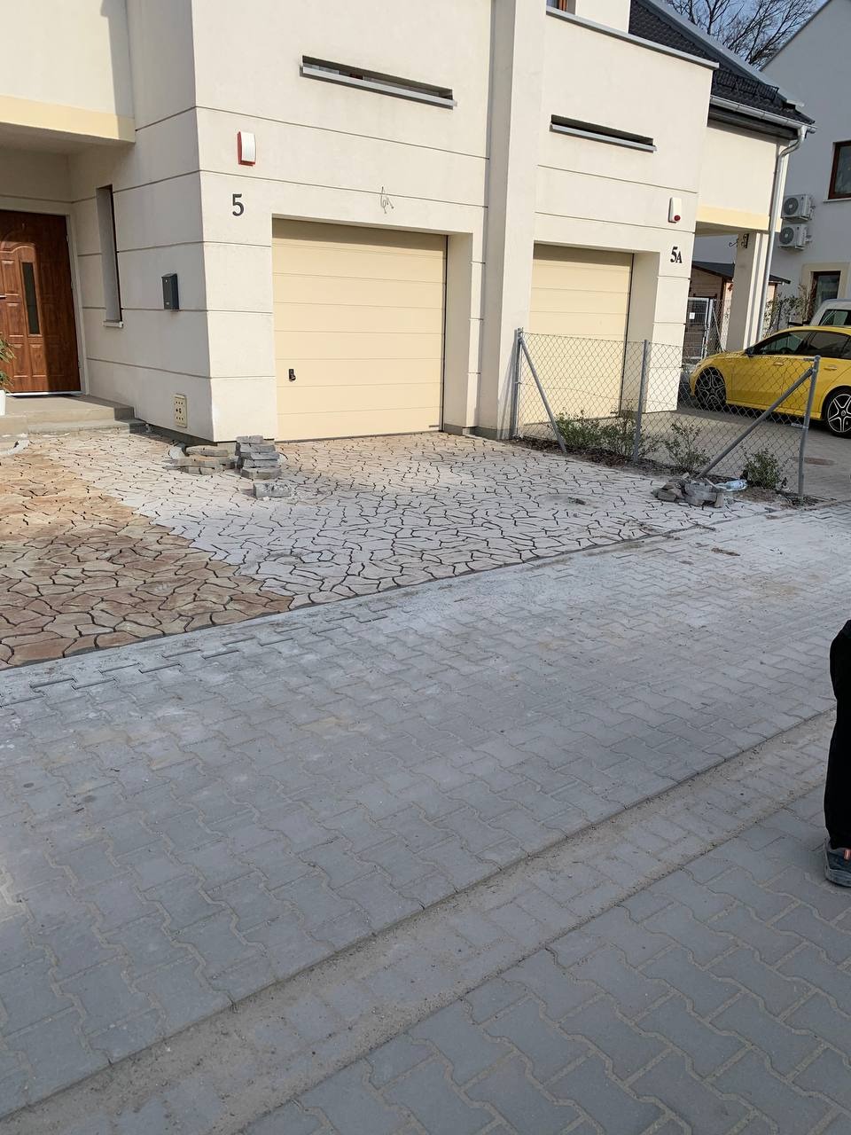 Układanie kostki brukowej przed domem z dwoma garażami, widoczne dwa rodzaje kostki: szara ułożona i brązowa w trakcie układania, obok sterta kostki do ułożenia.