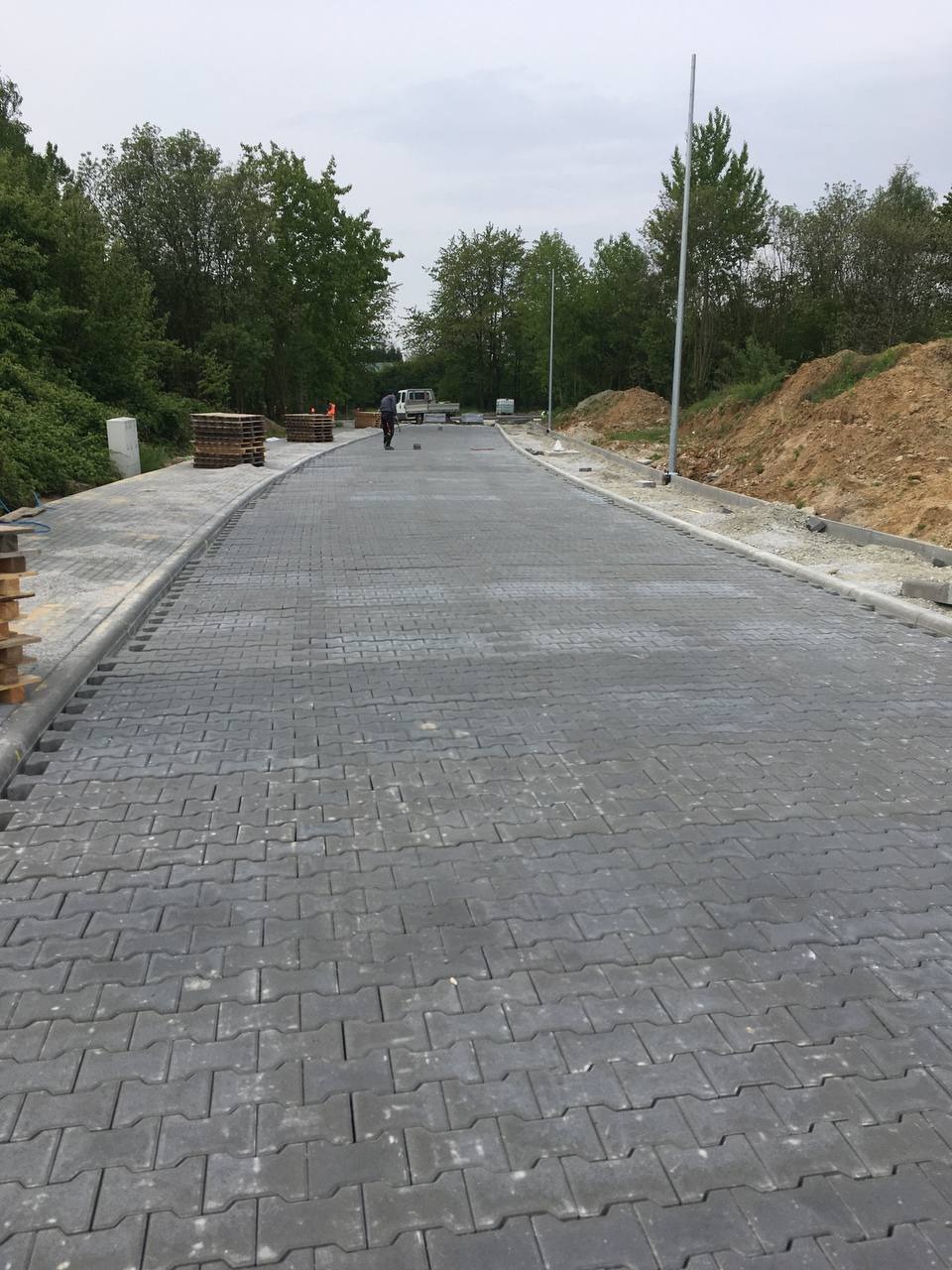 Nowo ułożona, szara kostka brukowa w kształcie litery 'H' na długiej drodze; w tle widać pracownika, ciężarówkę i stosy palet z kostką.