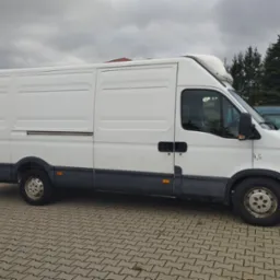 Biały furgon dostawczy Iveco z wysokim dachem, zaparkowany na kostce brukowej, widok z boku, pochmurne niebo w tle.