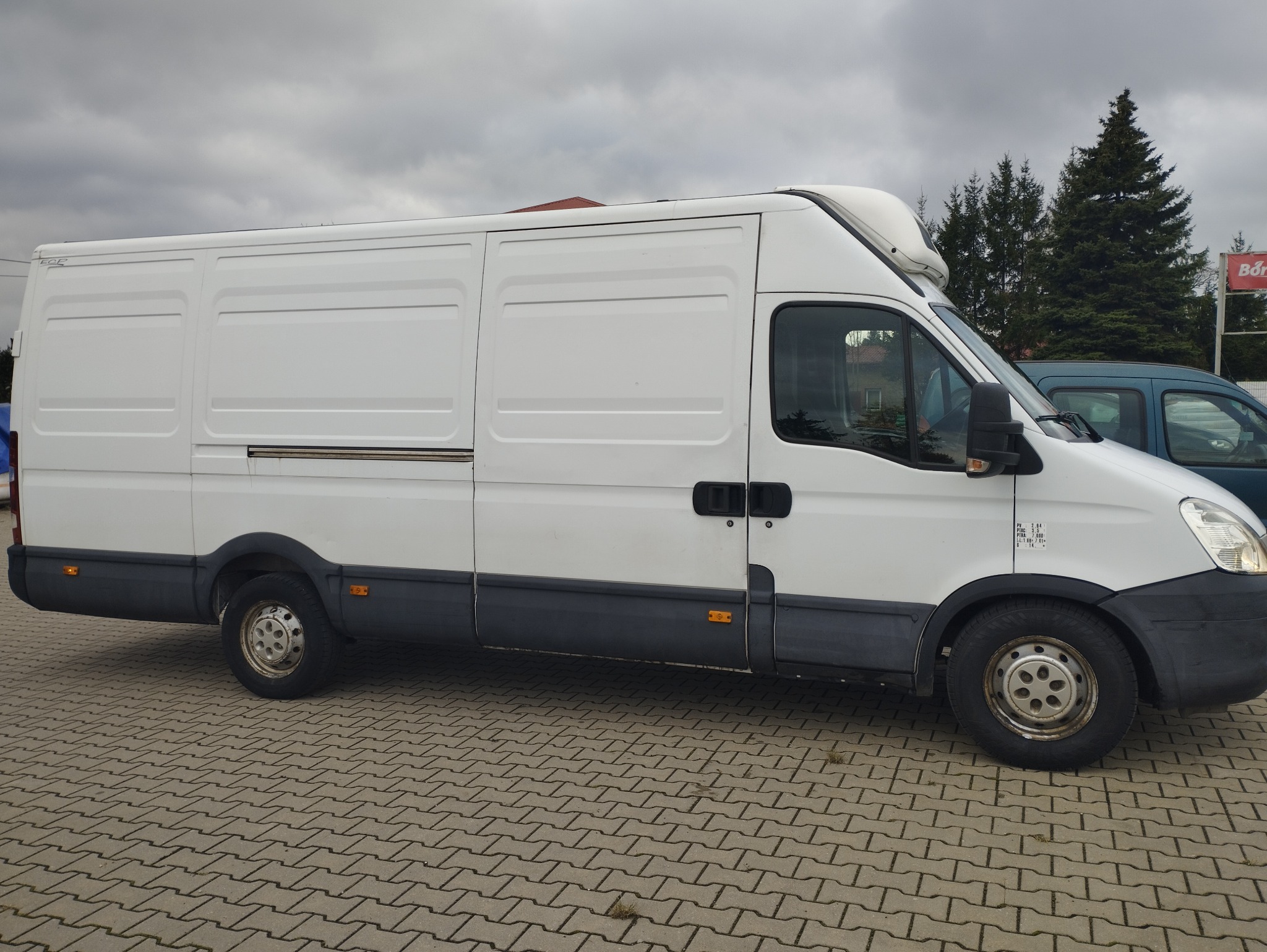 Biały furgon dostawczy Iveco z wysokim dachem, zaparkowany na kostce brukowej, widok z boku, pochmurne niebo w tle.