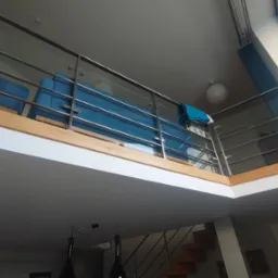 Wnętrze mieszkania z antresolą, widok z dołu na niebieską sofę za szklaną balustradą, w tle schody i okno dachowe.