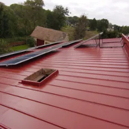 Czerwony dach z blachy trapezowej z zamontowanymi panelami fotowoltaicznymi i wyłazem dachowym, widok z góry na tle zieleni.