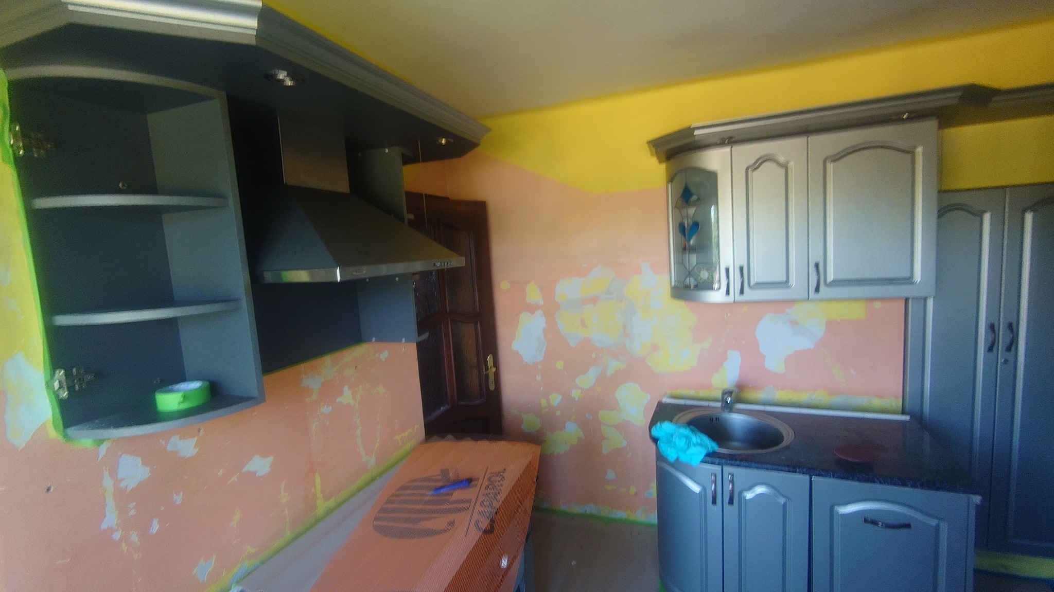 Kuchnia w trakcie malowania: widoczne szare szafki, okap, zlew, resztki starej farby na ścianach i karton z farbą Caparol.