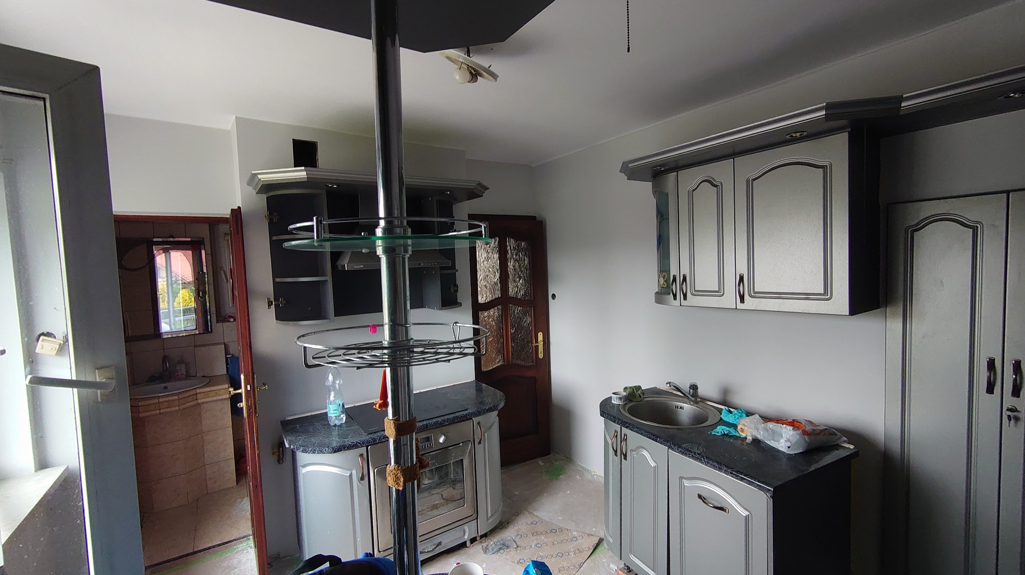 Szara kuchnia w trakcie remontu, ściany świeżo pomalowane na szaro, widoczne zabezpieczenia podłogi i niedokładności malowania przy podłodze, w tle otwarte drzwi do łazienki.