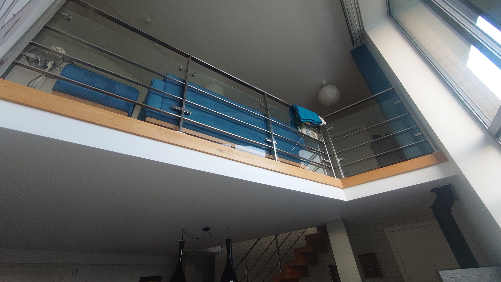 Wnętrze mieszkania z antresolą, widok z dołu na niebieską sofę za szklaną balustradą, w tle schody i okno dachowe.