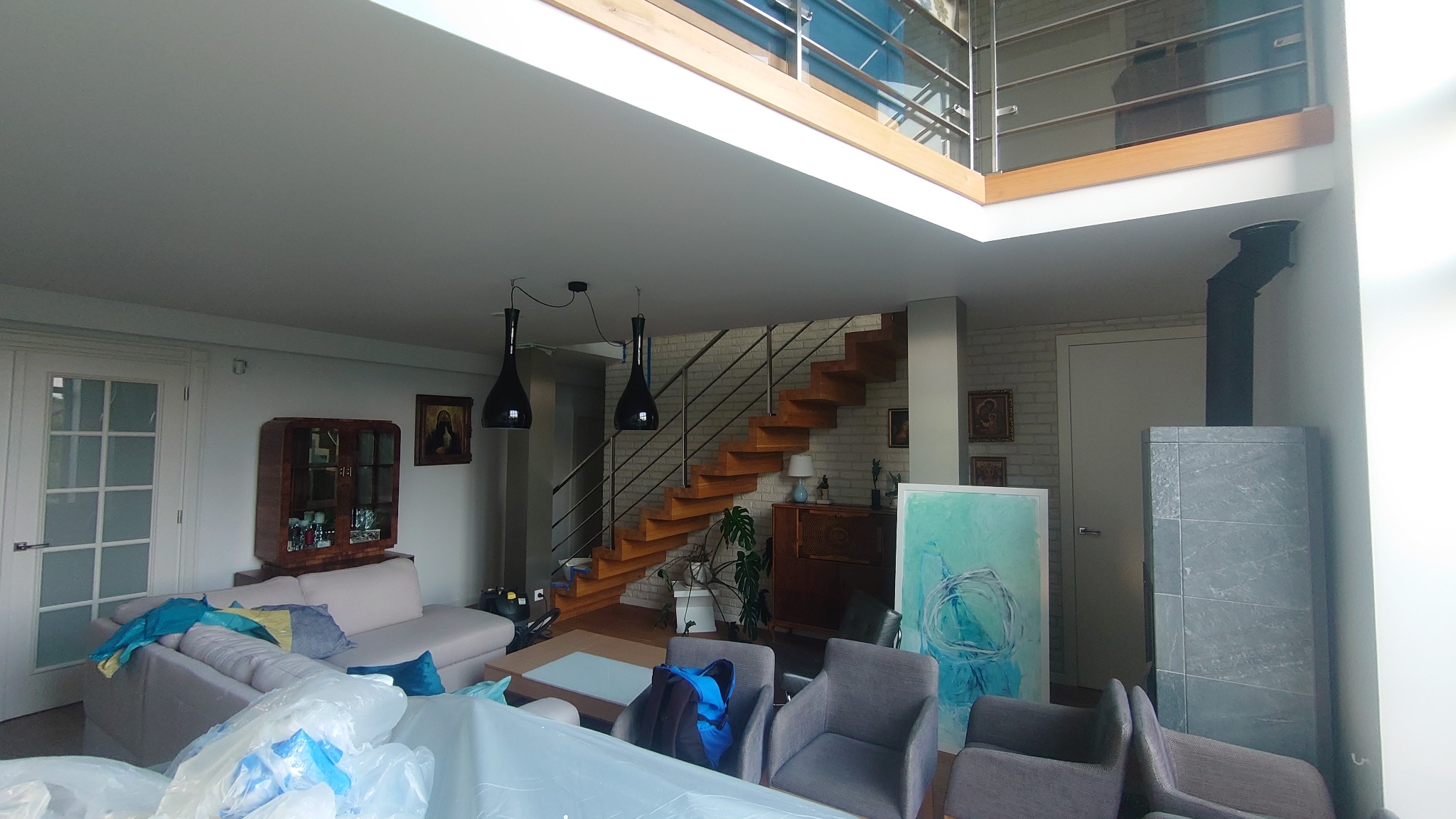 Salon w trakcie zabezpieczania przed remontem: meble przykryte folią ochronną, widoczne schody drewniane, nowoczesny piec kaflowy i wiszące lampy.