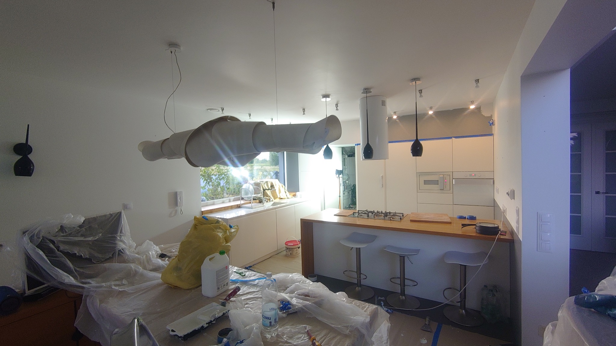 Kuchnia w trakcie malowania: zabezpieczone folią meble, widoczne taśmy malarskie na szafkach, wiadro z farbą i akcesoria malarskie na blacie.