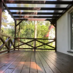 Zadaszony drewniany taras z balustradą krzyżową, widok na zieleń za siatką ogrodzeniową, brązowe deski podłogowe odbijają światło.