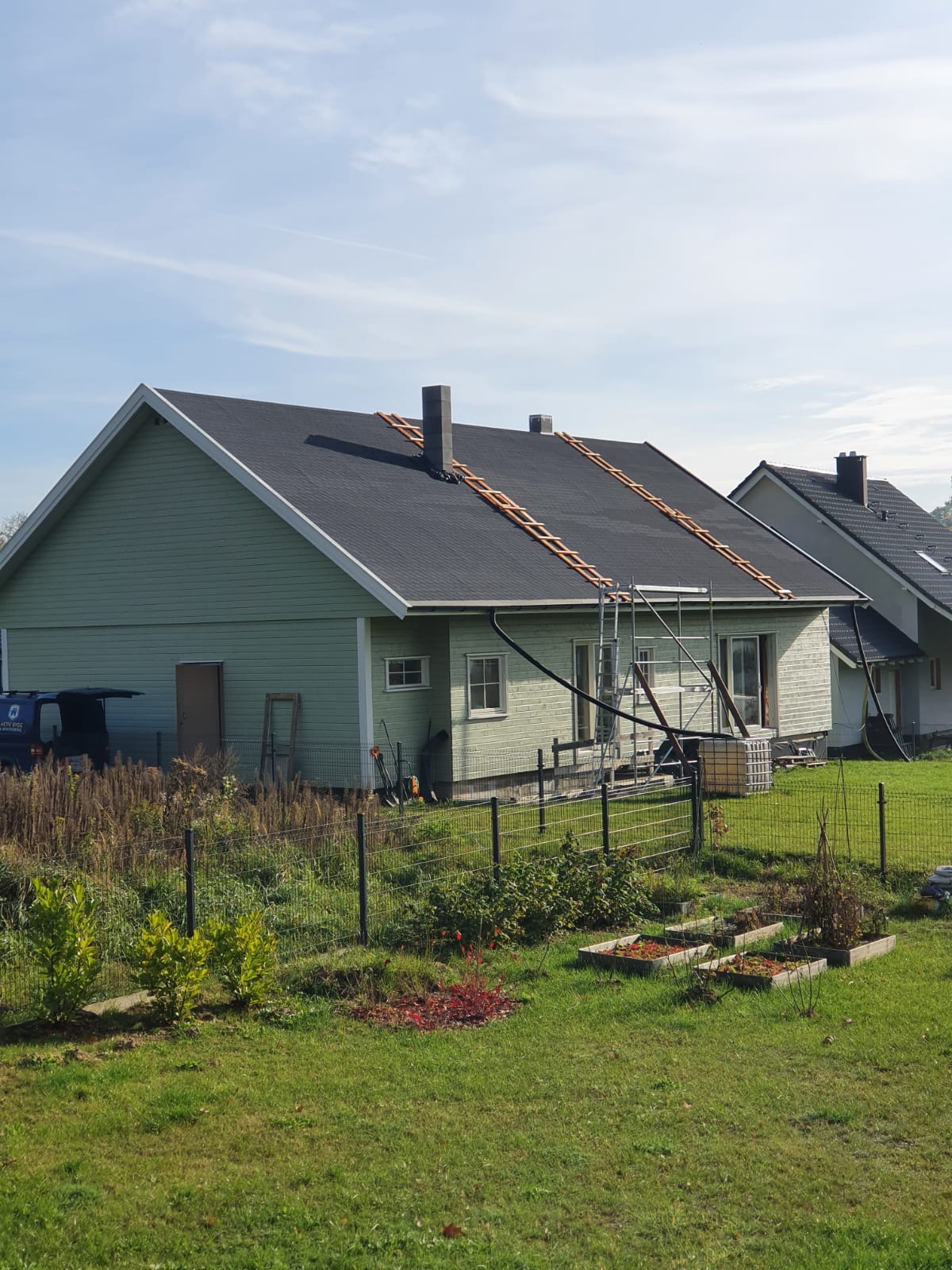 Jasnozielony dom w Obornikach Śląskich z czarnym dachem w trakcie budowy, widoczne drabiny dachowe i rusztowania, ogródek warzywny przed domem.