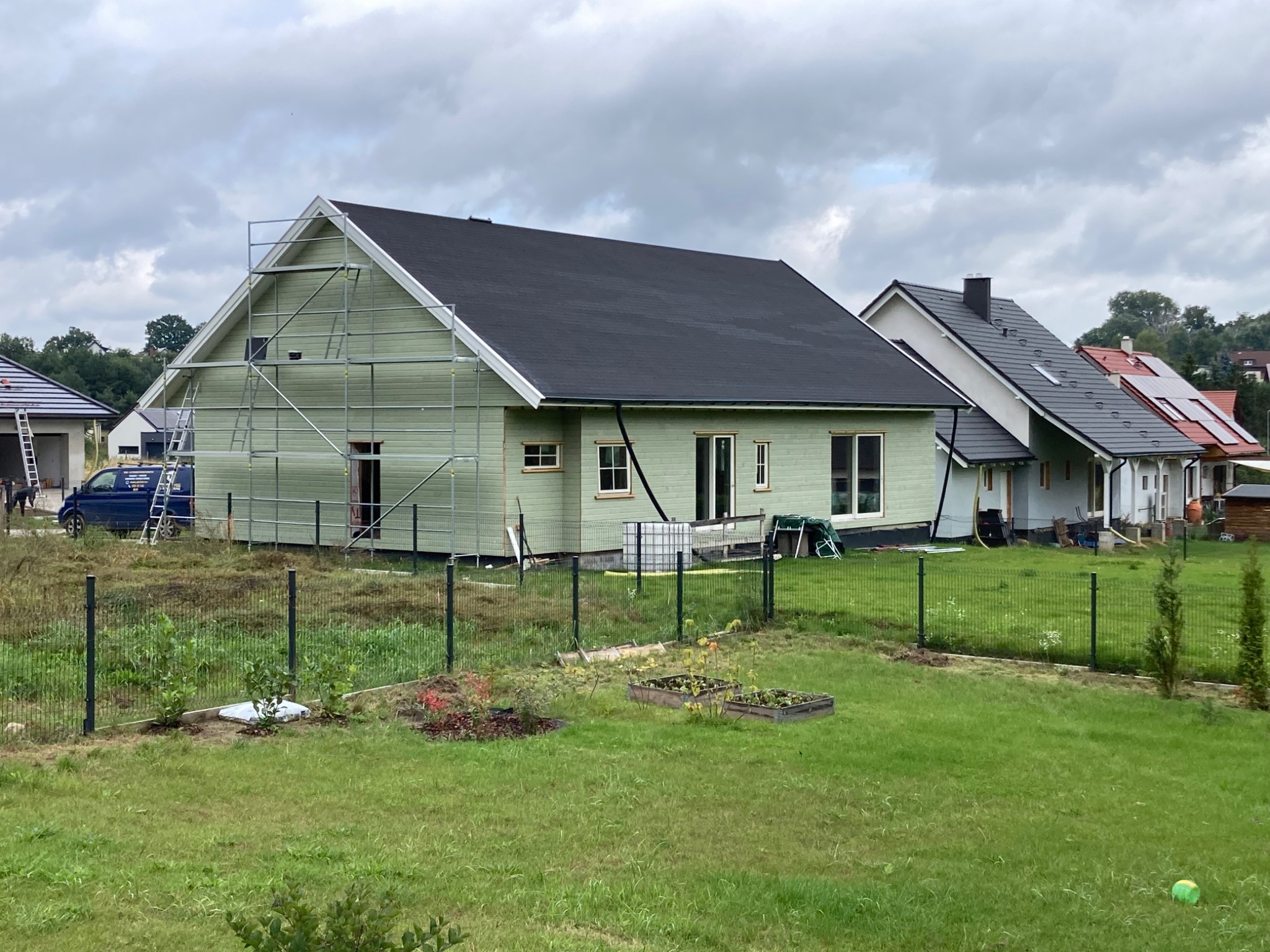 Budowa domu z drewnianą elewacją w trakcie realizacji, widoczne rusztowania, czarny dach i ogrodzenie posesji.