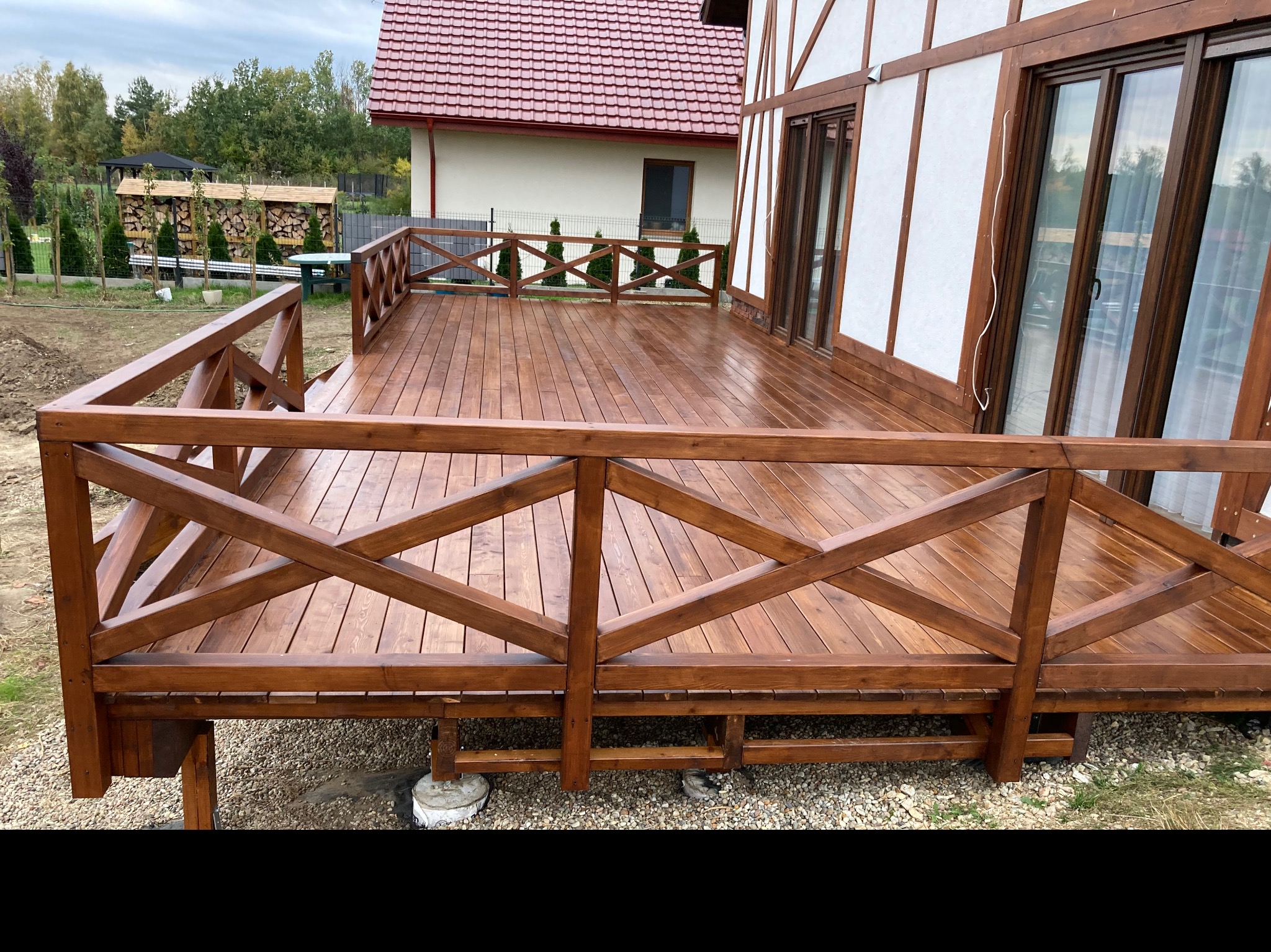 Drewniany taras z balustradą w stylu rustykalnym, przylegający do domu z elementami szkieletowymi, widok na ogród z przygotowanym drewnem opałowym.