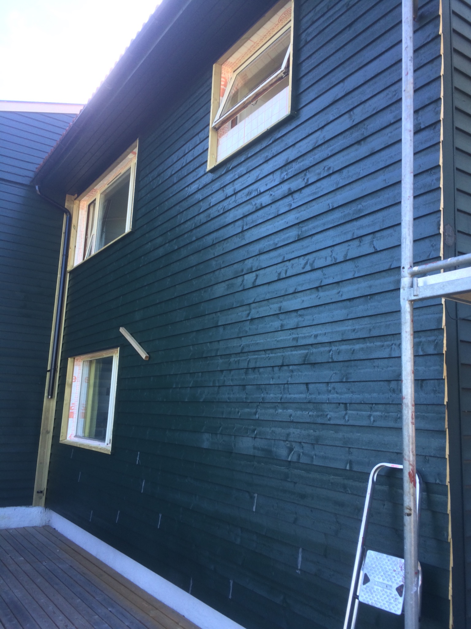 Ściana zewnętrzna budynku w trakcie budowy, pokryta ciemnozieloną drewnianą boazerią, okna w trakcie montażu, widoczne rusztowanie i drabina.