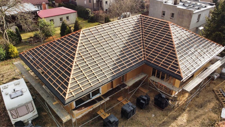 Budowa domu szkieletowego: widok z góry na konstrukcję dachu z łatami, z boku widoczny przyczepa kempingowa i rusztowania.