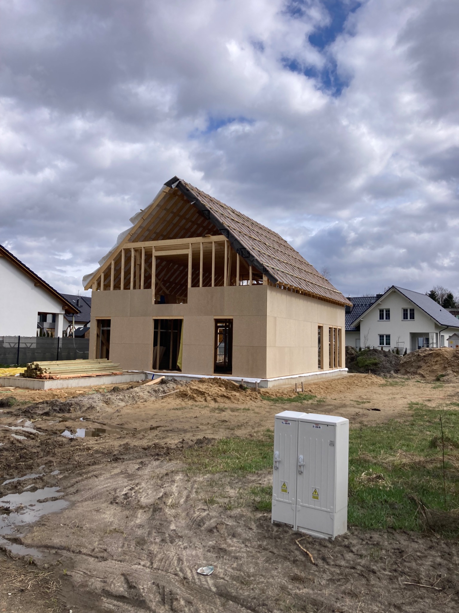 Surowa bryła domu w trakcie budowy z widoczną drewnianą konstrukcją dachu i ścian z płyt OSB, na tle pochmurnego nieba i sąsiednich domów.