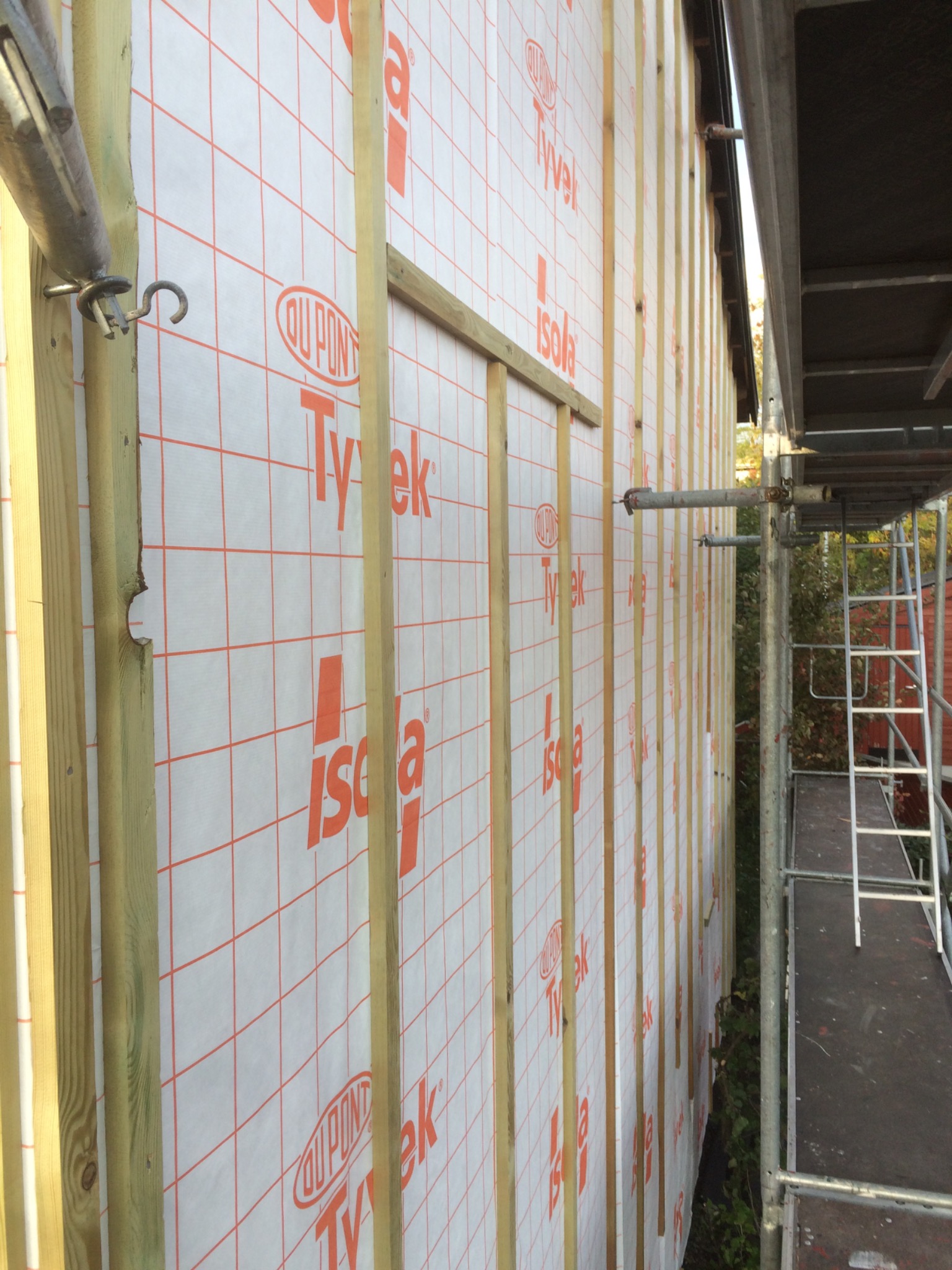 Ściana budynku w trakcie budowy, pokryta membraną izolacyjną z widocznymi łatami i elementami rusztowania.