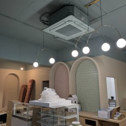 AEJ ENERGY Adam Jasina - Kasetonowa klimatyzacja sufitowa w lokalu usługowym, widoczne elementy instalacji i oświetlenia. Lokal w trakcie wykańczania, widoczne materiały i narzędzia.