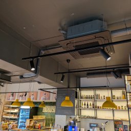 AEJ ENERGY Adam Jasina - Sufit lokalu gastronomicznego z zamontowaną kasetą klimatyzacyjną, widoczne rury wentylacyjne, oświetlenie punktowe i wiszące lampy. Ujęcie z dołu.