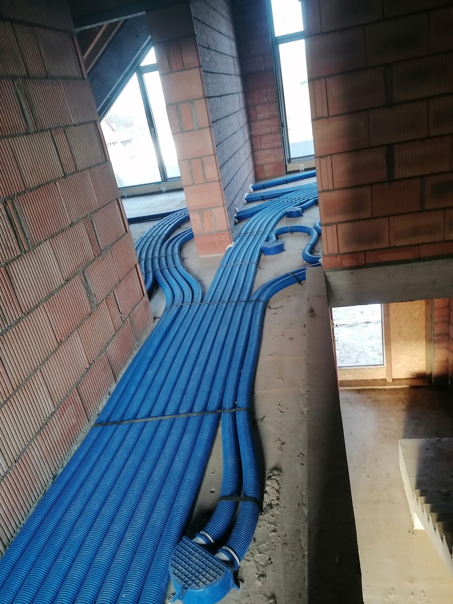 Niebieskie rury instalacji ogrzewania podłogowego ułożone w surowym wnętrzu budynku w trakcie budowy, widok z góry, ceglane ściany.