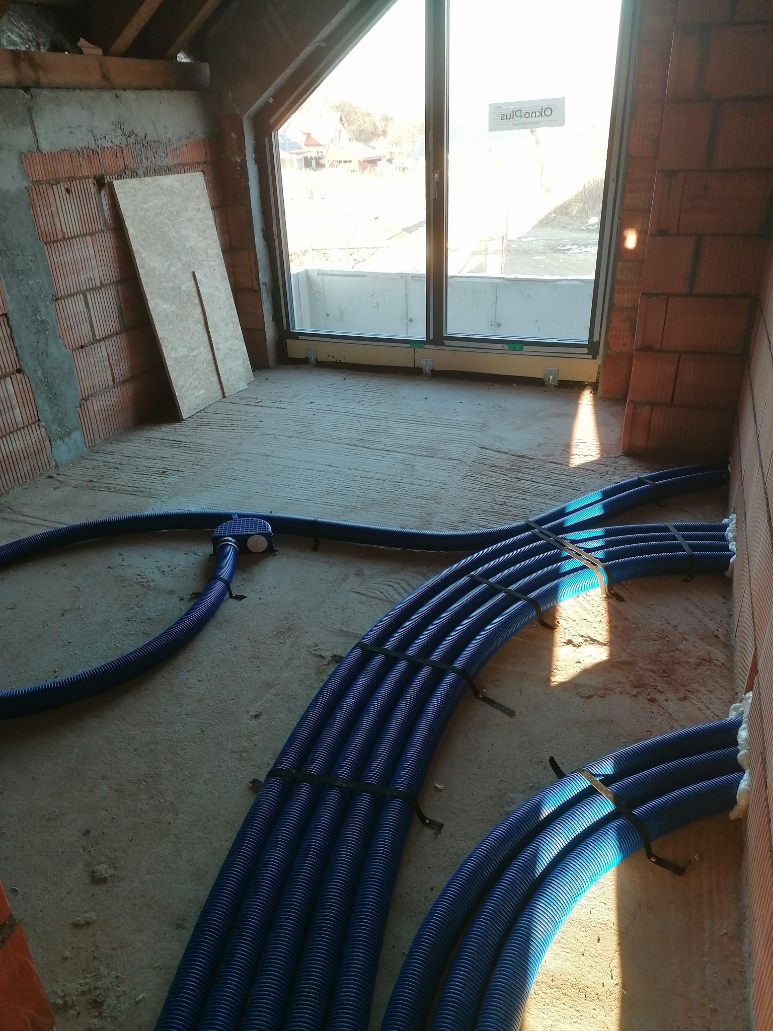 Niebieskie rury instalacji ogrzewania podłogowego ułożone w surowym wnętrzu z oknem. Widoczne mocowania i fragmenty ścian z cegły. Etap prac przed wylaniem posadzki.